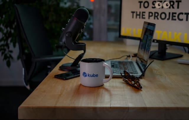 Kube-branded coffee mug beside a laptop and podcast microphone on a wooden desk.